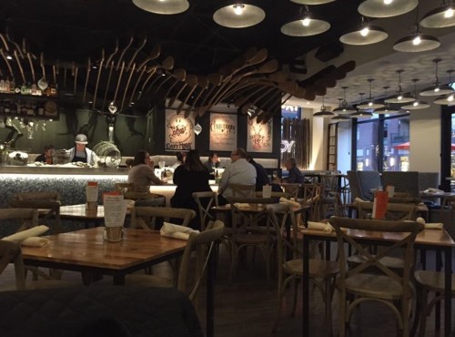 A cozy restaurant interior with wooden tables, chairs, and a bar area, featuring modern decor and dim lighting.