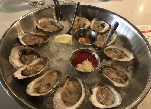 A metal platter filled with fresh oysters on ice, accompanied by lemon wedges and dipping sauces.