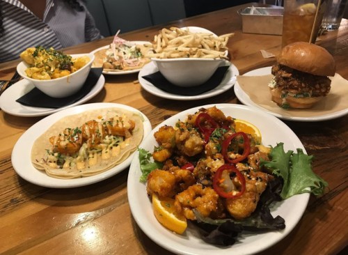A table with various dishes: tacos, fried chicken sandwich, fries, and cauliflower bites garnished with peppers and herbs.