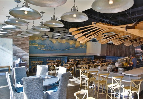 Bright restaurant interior with wooden decor, blue seating, and unique light fixtures, featuring a large wall mural.