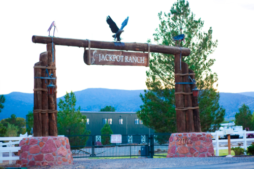 Wooden entrance gate to Jackpot Ranch, featuring an eagle and decorative elements, surrounded by trees and mountains.