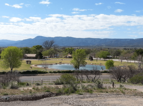 A scenic view of a green park with a pond, surrounded by trees and mountains under a partly cloudy sky.