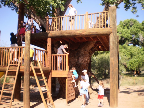 Children and adults play on a large wooden treehouse with ladders and a climbing wall under a clear blue sky.