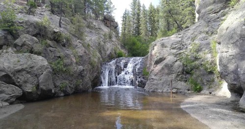 A serene waterfall cascades into a calm pool, surrounded by rocky cliffs and lush greenery in a forested area.