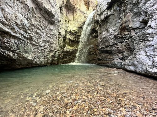 A serene waterfall cascades into a clear pool surrounded by rocky cliffs and pebbled ground.