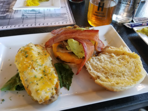 A plate with a cheesy baked potato, a burger with bacon and guacamole, and a toasted bun on the side.