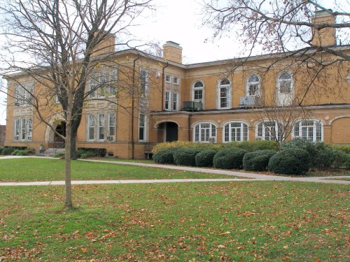 A historic brick building with large windows, surrounded by grass and trees, on a cloudy day.