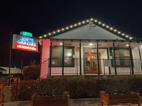 A brightly lit restaurant at night with a neon sign reading "Joe L. Garcia's Mexican Dishes" and festive outdoor decor.