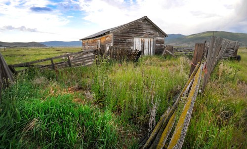 Abandoned wooden barn surrounded by overgrown grass and a dilapidated fence under a cloudy sky.