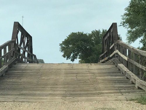 A wooden bridge with a gravel approach, surrounded by trees and a cloudy sky.