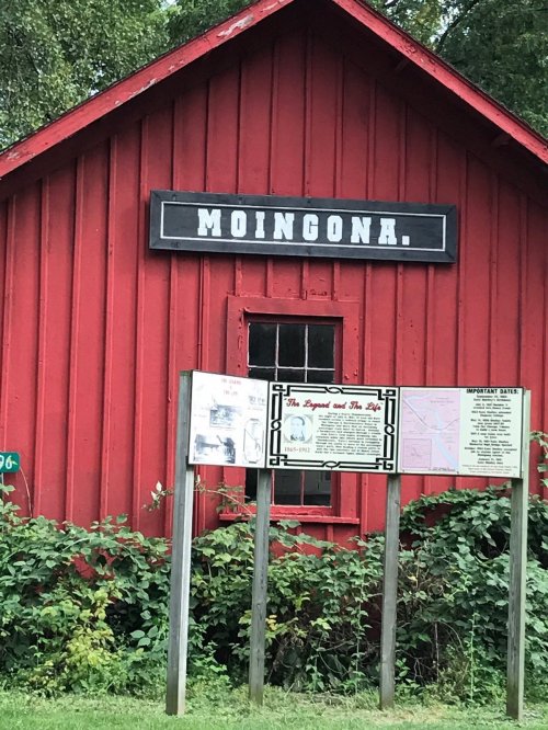 A red wooden building with a sign reading "MOINGONA" and an informational board in front, surrounded by greenery.