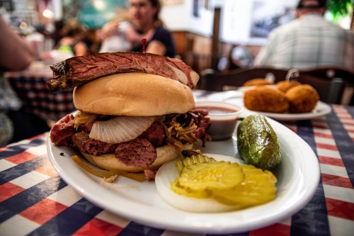 A hearty sandwich piled with meats, onions, and pickles, served with a side of pickles and cornbread on a checkered tablecloth.