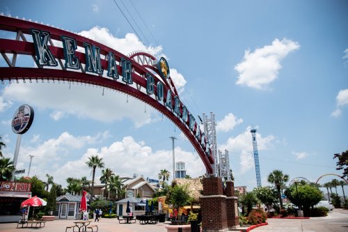 Entrance archway of Kenah Boardwalk with blue skies, palm trees, and amusement rides in the background.
