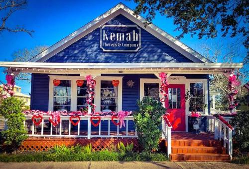 A charming blue house with a red door, decorated with pink hearts and flowers, labeled "Kemah Flowers & Company."