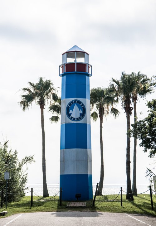 A colorful lighthouse stands among palm trees, overlooking the ocean under a cloudy sky.