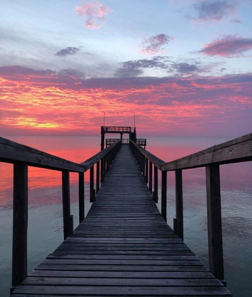 A wooden pier extends into calm waters, framed by a vibrant sunset with pink and orange hues in the sky.