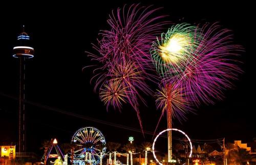 Colorful fireworks illuminate the night sky above a carnival with a Ferris wheel and a tall observation tower.