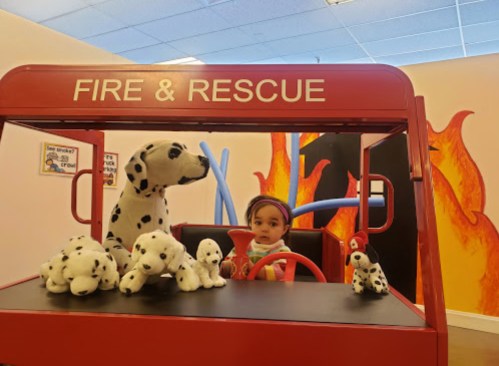 A child sits in a toy fire truck surrounded by plush dalmatian puppies in a colorful play area.