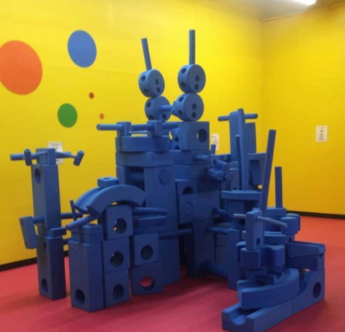 A colorful playroom with a large structure made of blue building blocks against a bright yellow wall.
