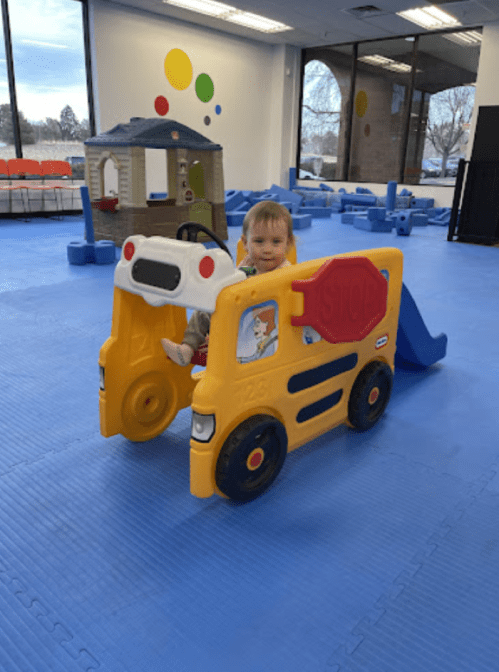 A toddler sits in a colorful toy bus in a playroom with blue flooring and various play structures in the background.