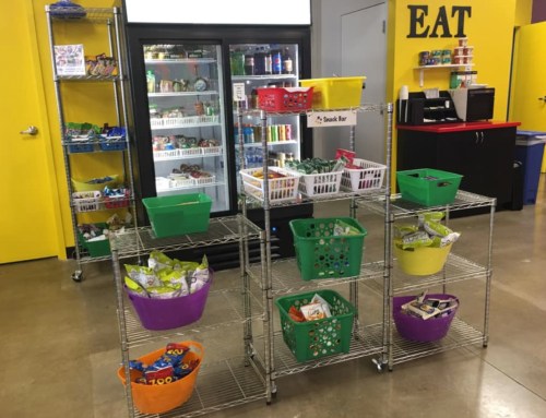 A colorful snack bar with shelves and baskets filled with various snacks, next to a refrigerator and a bright yellow wall.
