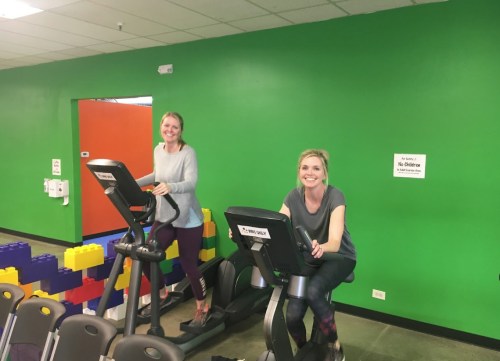 Two women are smiling while exercising on elliptical machines in a brightly colored gym.