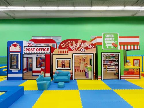 Colorful play area featuring a post office, restaurant, and supermarket with a blue couch and bright flooring.