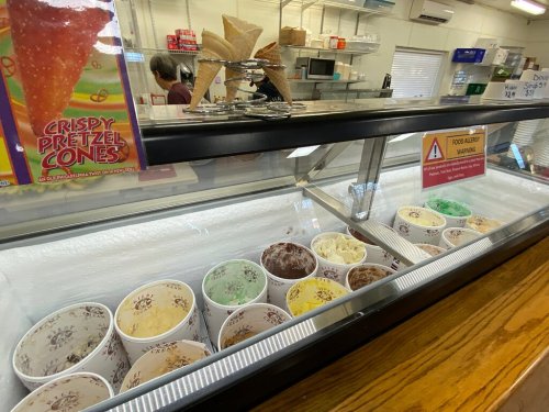 A display case filled with various ice cream flavors in cups, with a sign for crispy pretzel cones in the background.