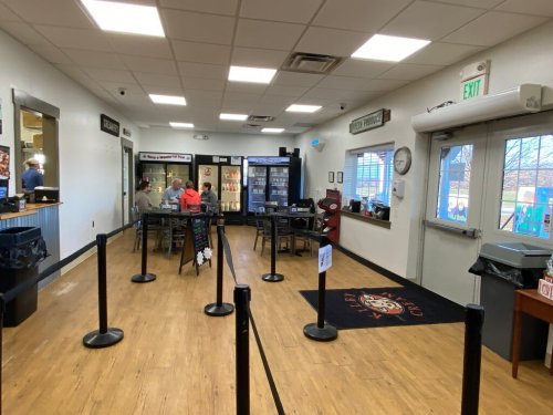 Interior of a casual dining space with tables, a drink cooler, and a waiting area marked by stanchions.