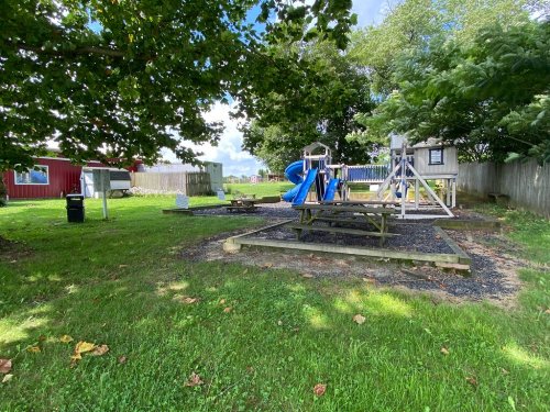 A playground with a slide and swings, surrounded by grass and trees, in a sunny outdoor setting.