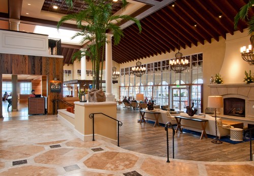 A spacious hotel lobby with high ceilings, elegant decor, and palm trees, featuring seating areas and a reception desk.