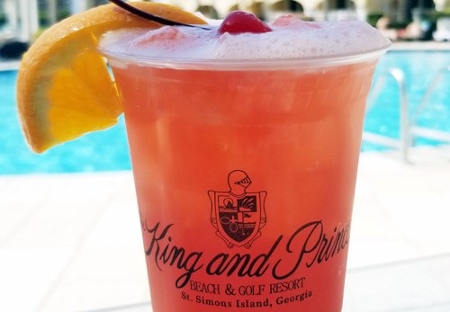 A refreshing cocktail in a clear cup, garnished with a cherry and orange slice, with a pool in the background.