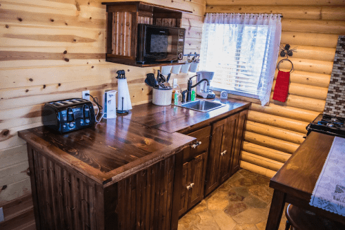 Cozy wooden kitchen with a countertop, microwave, toaster, and window, featuring rustic log walls and stone flooring.