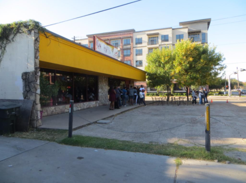 A restaurant with a yellow awning and a line of people outside, surrounded by trees and an apartment building in the background.