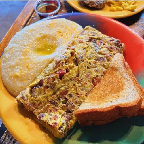 A colorful plate with a fluffy omelet, grits with butter, and two slices of toasted bread.