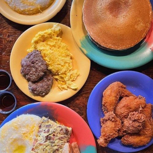 A colorful spread of breakfast plates featuring pancakes, eggs, sausage, grits, and fried chicken.