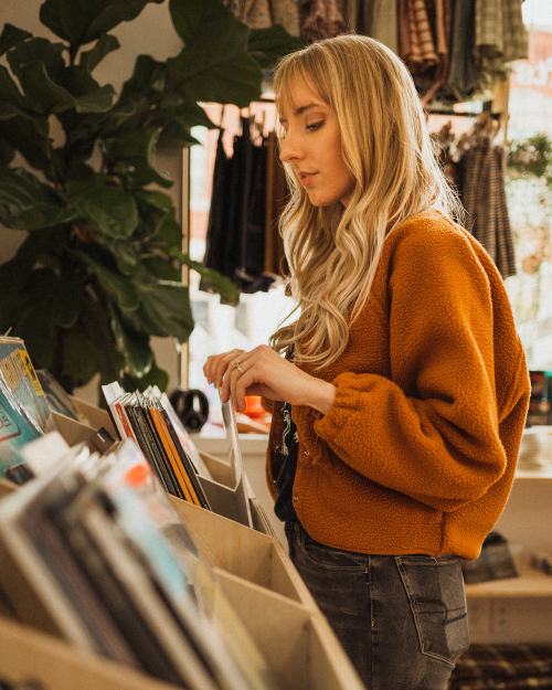 A woman with long blonde hair browses through vinyl records in a cozy shop filled with plants and clothing.