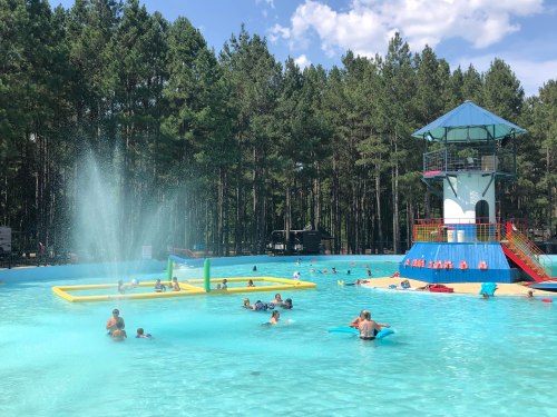 A vibrant swimming pool surrounded by trees, with people enjoying the water and a lifeguard tower in the background.