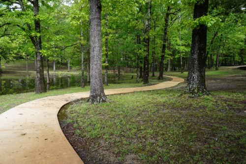 A winding path through a lush green forest, bordered by trees and a calm body of water nearby.