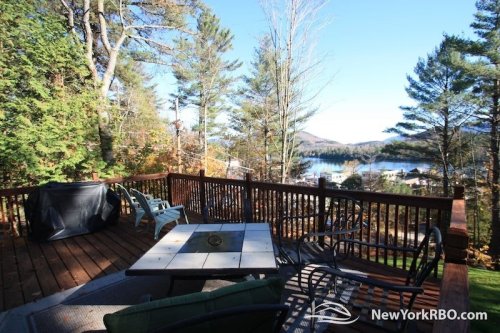 A wooden deck with outdoor furniture overlooks a serene lake surrounded by trees and mountains.