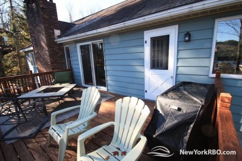 A wooden deck with two white chairs, a grill, and a door leading into a blue house. Trees are visible in the background.