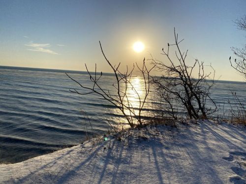 A serene winter scene of a sun setting over calm waters, with bare branches and snow-covered ground in the foreground.
