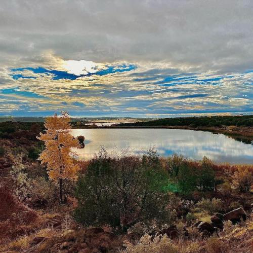 A serene landscape featuring a calm lake, surrounded by trees and colorful autumn foliage under a cloudy sky.