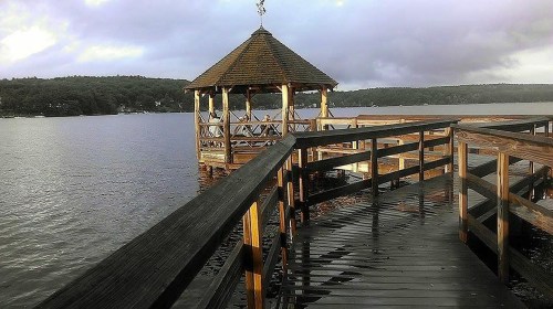 A wooden pier leads to a gazebo on a calm lake, surrounded by trees under a cloudy sky.