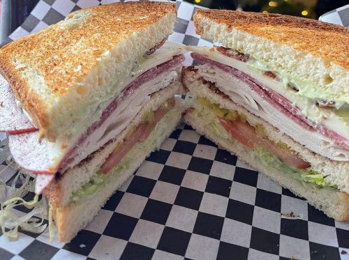 A close-up of a toasted sandwich with layers of turkey, ham, lettuce, tomato, and a creamy spread, served on checkered paper.