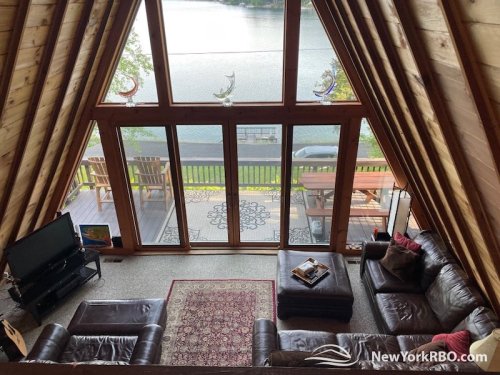 Cozy A-frame cabin interior with large windows overlooking a lake, featuring a living area and wooden deck.