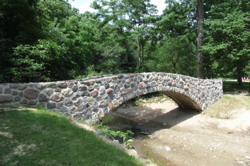 A stone arch bridge over a dry creek, surrounded by lush green trees and grass in a park setting.