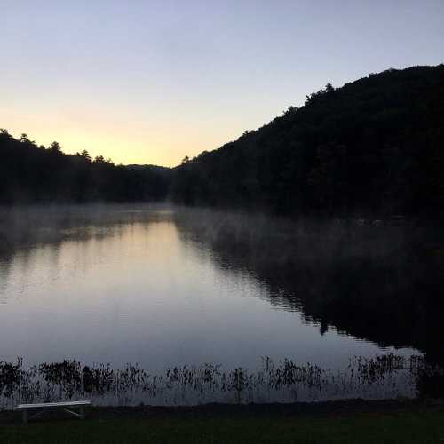 A serene lake at dawn, surrounded by trees, with mist rising from the water and a soft golden light in the sky.