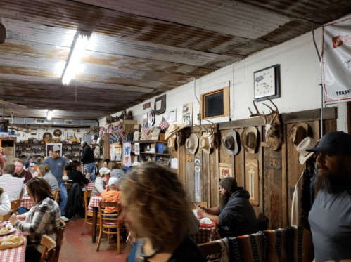 A rustic restaurant interior with patrons dining, decorated with hats, antlers, and vintage signs on the walls.
