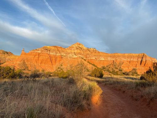 A scenic view of red rock formations under a cloudy sky, with a dirt path and grassy foreground.